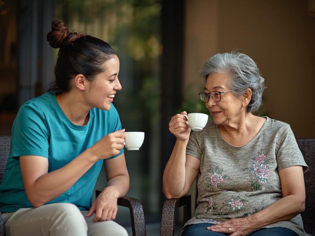 a carer wearing a teal color plain tshirt drinking tea with a cancer patient in home. both are sitting on chairs outdoor